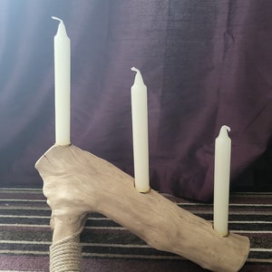May include: A rustic wooden candle holder with three candle slots. The holder is made from a natural branch of wood and has a rope wrapped around the base. Three white candles are placed in the slots.