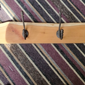 May include: A wooden coat rack with four metal hooks. The rack is made of natural wood with a live edge and has a light brown colour. The hooks are black metal and have a simple design.