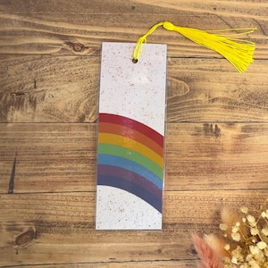 May include: A white bookmark with a rainbow design and a yellow tassel. The bookmark has a speckled white background.