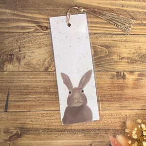 May include: A white bookmark with a brown rabbit illustration. The bookmark has a pink glitter background and a gold tassel.