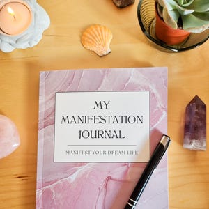 May include: A pink and white marble-patterned journal with the text "MY MANIFESTATION JOURNAL" and "MANIFEST YOUR DREAM LIFE" on the cover. A black pen is lying on top of the journal.