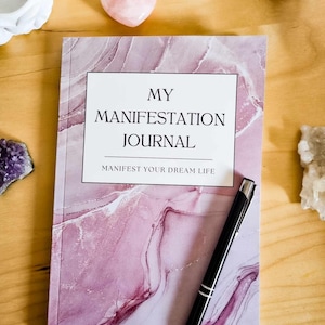 May include: A pink marble-patterned journal with the text "MY MANIFESTATION JOURNAL" and "MANIFEST YOUR DREAM LIFE" on the front cover. A black pen is lying on the journal.