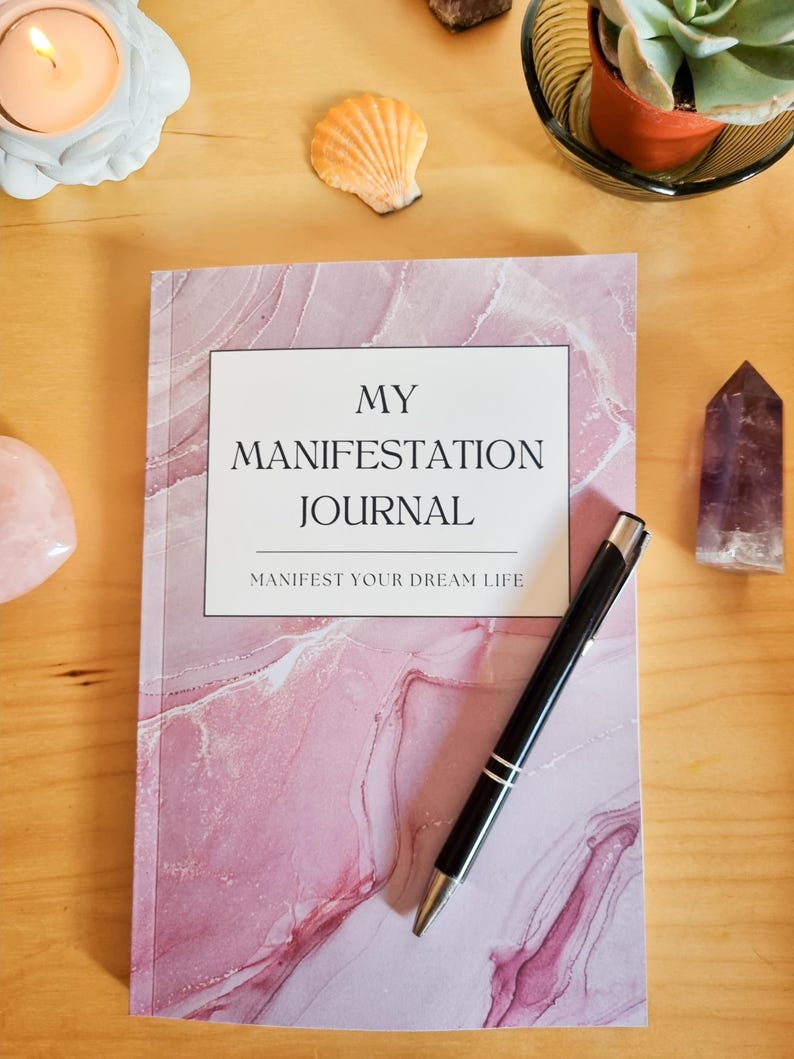May include: A pink and white marble-patterned journal with the text "MY MANIFESTATION JOURNAL" and "MANIFEST YOUR DREAM LIFE" on the cover. A black pen is lying on top of the journal.