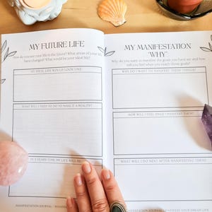 May include: An open journal with the title "My Future Life" and "My Manifestation 'Why'" on the pages. The journal has prompts for reflection and goal setting. The journal is on a wooden surface with a pink crystal, a seashell, and a small potted succulent.