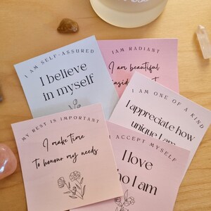 May include: Five pink and blue affirmation cards with positive messages about self-love, self-acceptance, and self-care. The cards feature simple floral illustrations and the text "I am self-assured, I believe in myself", "My rest is important, I make time to honour my needs", "I am radiant, I am beautiful inside and out", "I am one of a kind, I appreciate how unique I am", and "I accept myself, I love who I am".