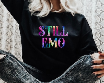 May include: A black sweatshirt with the words "STILL EMO" printed on the front in rainbow colours. The sweatshirt is being worn by a person sitting down with their legs crossed. The person is wearing ripped jeans and leopard print trainers.
