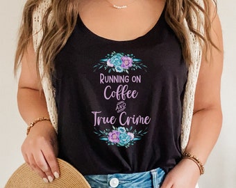May include: A black tank top with the text "Running on Coffee and True Crime" in purple and blue lettering with floral accents. The model is wearing denim shorts and a white cardigan.