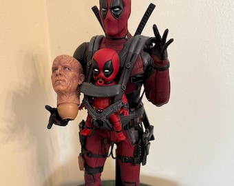 Può includere: Una action figure di Deadpool rossa e nera che tiene una action figure di Deadpool bambino in un marsupio. La figura tiene una testa mozzata nell'altra mano. La figura è in piedi su una base nera.