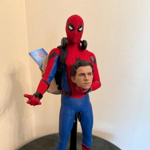 May include: A Spider-Man action figure in a red and blue suit, with a backpack and headphones. The figure is holding a separate head. The figure is standing on a black stand.