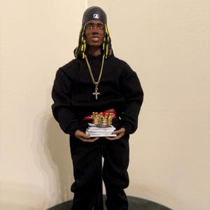 Puede incluir: Muñeca vestida con una sudadera con capucha negra y pantalones a juego, con una gorra de béisbol y un collar de cruz dorada. La muñeca sostiene una pila de papeles y una corona dorada. La muñeca lleva zapatillas deportivas negras, doradas y blancas.