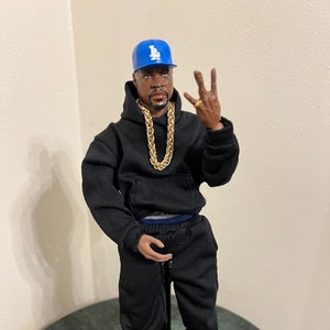 May include: A black action figure wearing a black hoodie, black pants, and blue and white sneakers. The figure is wearing a gold chain necklace and a blue baseball cap with the letters "LA" on it. The figure is standing on a black stand.