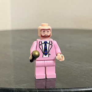 May include: A pink Lego minifigure wearing a pink suit with a blue and white striped tie and holding a gold microphone.
