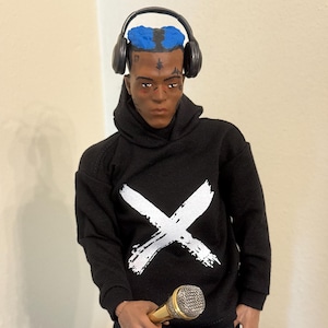 May include: A black action figure wearing a black hoodie with a white "X" design, black pants with white "X" designs, and black headphones. The figure has blue hair and holds a gold microphone.