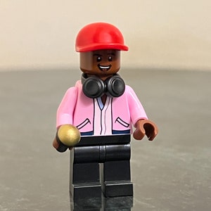 Puede incluir: Una minifigura de Lego con una chaqueta rosa, auriculares negros, una gorra de béisbol roja y sosteniendo un micrófono dorado.
