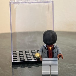 May include: A gray and white Lego minifigure holding a gold microphone. The minifigure is wearing a gray jacket, a red shirt, and white pants. The minifigure is standing in a clear plastic display case with a black base.