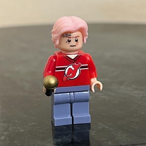 May include: A pink-haired Lego minifigure wearing a red jersey with a white and black logo, blue jeans, and holding a gold microphone.