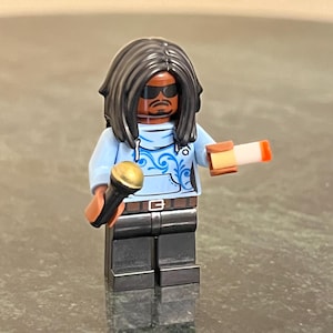 May include: A Lego minifigure with black hair, a blue hoodie with a floral design, black pants, and sunglasses. The minifigure is holding a microphone in one hand and a small white rectangle in the other.