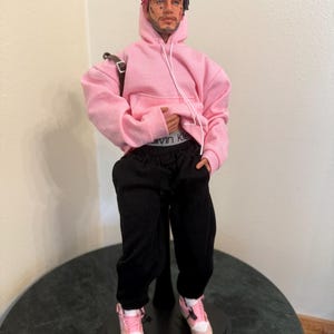 Puede incluir: Muñeca de una persona con sudadera rosa, pantalón de chándal negro y zapatillas rosas y blancas. La muñeca tiene una mochila y un peinado bicolor, rosa y negro. La muñeca está sobre un soporte negro.