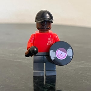 May include: A red Lego minifigure with a black hat and a black mustache. The minifigure is holding a microphone in one hand and a black vinyl record in the other. The minifigure is wearing a red shirt with the text "I FEEL LIKE PABLO" printed on it.