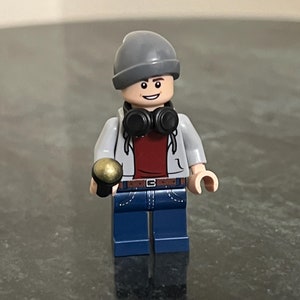 May include: A gray and white Lego minifigure wearing a gray beanie, a red shirt, a gray jacket, blue jeans, and black headphones. The minifigure is holding a gold microphone.