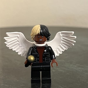 May include: A black and white Lego minifigure with white wings, a black leather jacket, a red shirt, and black pants. The minifigure is holding a gold microphone.
