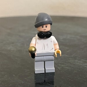 Op de afbeelding: Een grijs en wit Lego minifiguur met een grijze muts en zwarte bril, die een gouden microfoon vasthoudt.