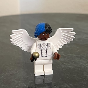 May include: A white Lego figurine with wings, wearing a white suit with a black bow tie and holding a gold microphone. The figurine has black hair with blue highlights.