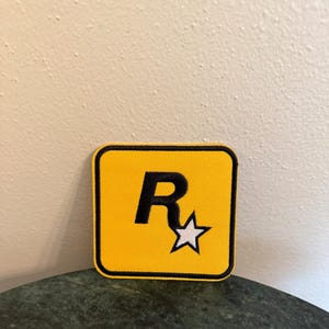 May include: A yellow square patch with a black border and a black letter "R" with a white star inside. The patch is on a green marble surface.