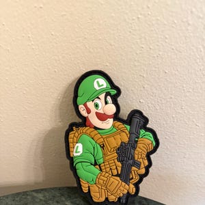 May include: A green and brown embroidered patch featuring a cartoon character wearing a green hat with an 'L' on it, a green shirt, and a brown tactical vest. The character is holding a black rifle.