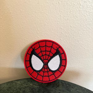May include: A round, embroidered patch featuring the iconic mask of Spider-Man. The patch has a red background with a black web design and white eye shapes. The patch is a classic superhero accessory.