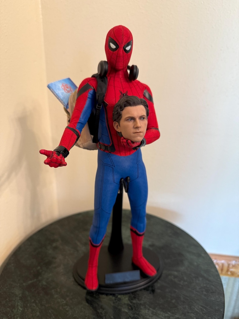 May include: A Spider-Man action figure in a red and blue suit, with a backpack and headphones. The figure is holding a separate head. The figure is standing on a black stand.