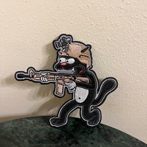 May include: A black cat wearing a tan helmet and holding a rifle. The cat is embroidered on a patch.