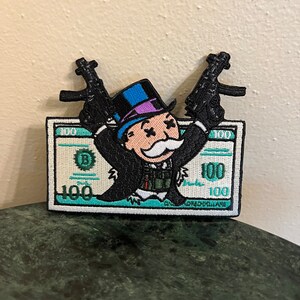 May include: Embroidered patch featuring a cartoon Monopoly Man holding two guns, standing on a $100 bill. The Monopoly Man is wearing a top hat, a monocle, and a mustache.