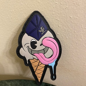 Può includere: Un cono gelato di cartone animato con un viso sorridente che indossa un cappello da pilota blu con un distintivo alato. Il gelato è rosa e bianco con una goccia blu. Il cono è marrone e bianco.