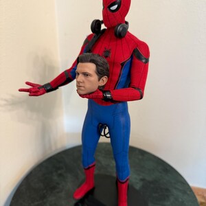 May include: A Spider-Man action figure in a red and blue suit, with a removable head. The figure has a detailed design, including a mask, web-shooters, and headphones. The figure is standing on a black base.