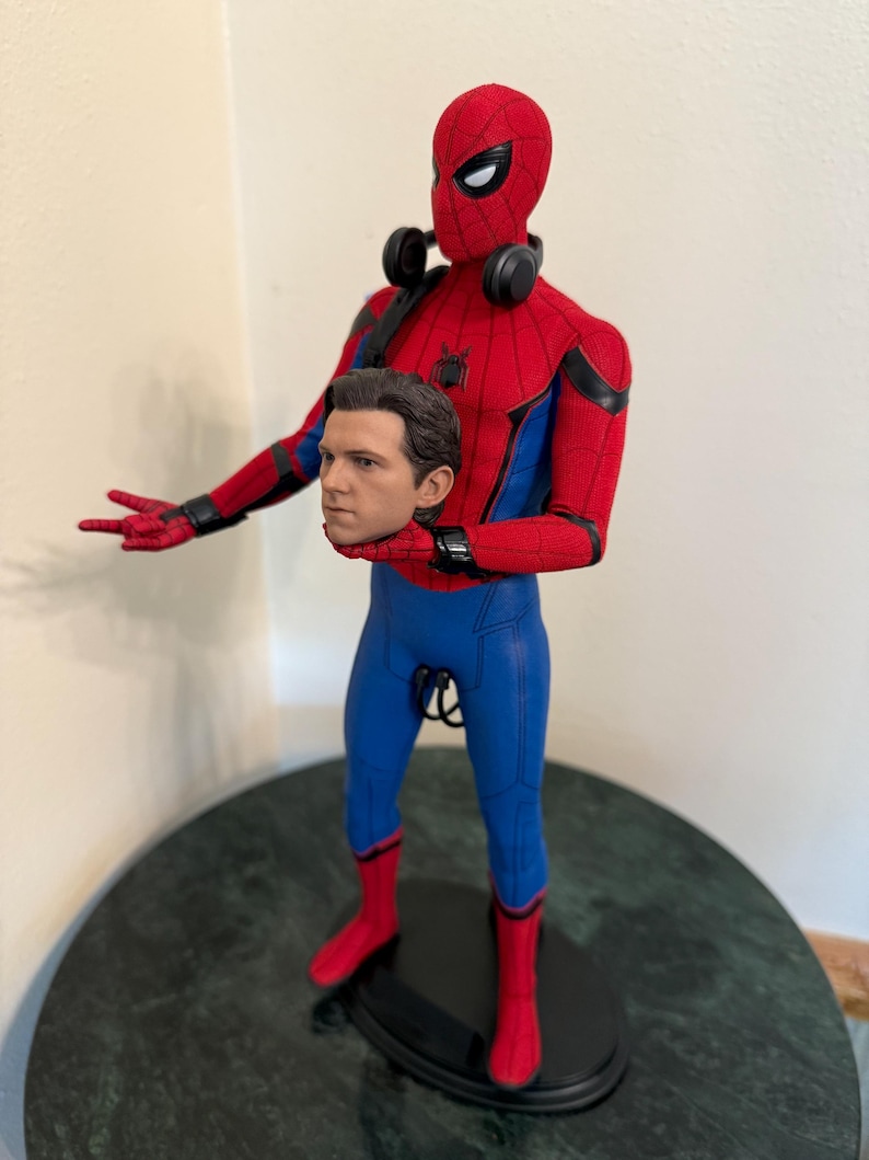 May include: A Spider-Man action figure in a red and blue suit, with a removable head. The figure has a detailed design, including a mask, web-shooters, and headphones. The figure is standing on a black base.