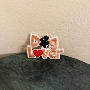 Pode incluir: Um remendo branco bordado com as palavras "Dog Lover" em fio laranja. Uma pegada de pata preta está no meio do "D" e um coração vermelho está no meio do "o".