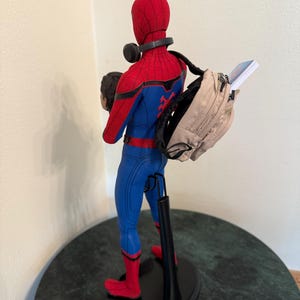 May include: A Spider-Man action figure in a red and blue suit, with a backpack and headphones. The figure is on a black stand, with a head in its left hand. The backpack is tan and has a book sticking out.