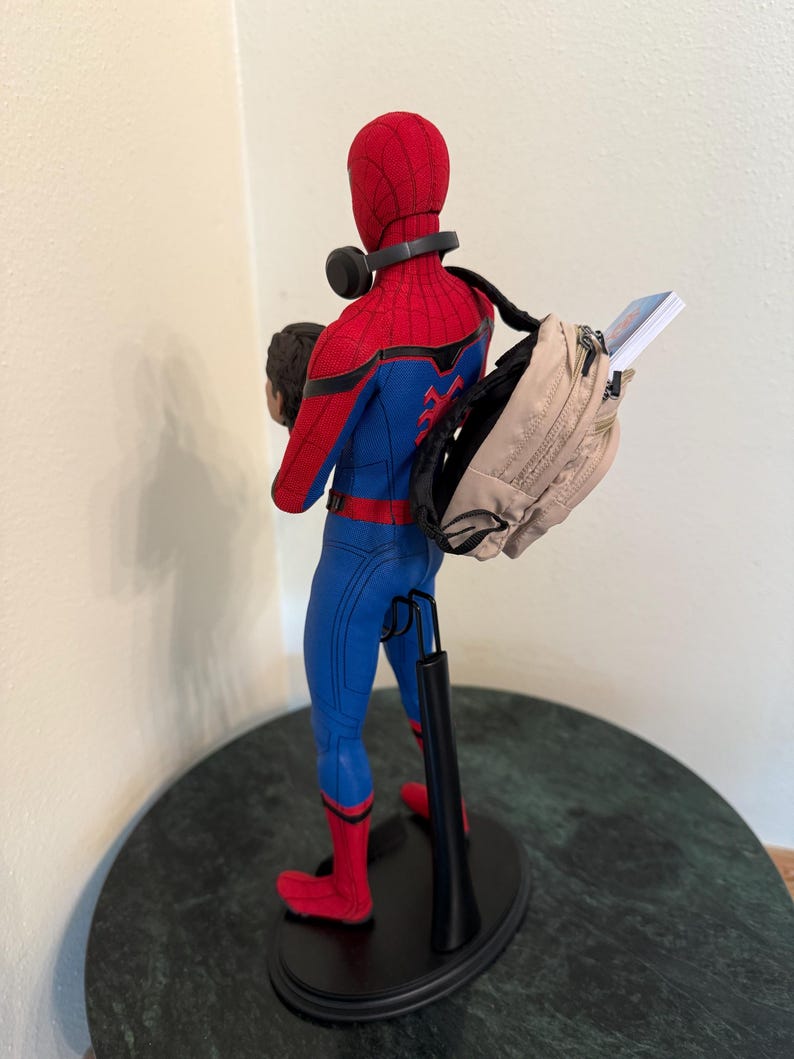 May include: A Spider-Man action figure in a red and blue suit, with a backpack and headphones. The figure is on a black stand, with a head in its left hand. The backpack is tan and has a book sticking out.
