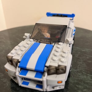 May include: A grey and blue Lego car with a blue stripe down the bonnet and a Lego figure in the driver's seat.