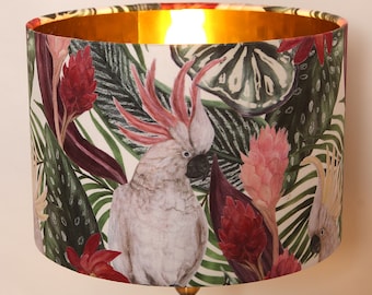May include: A white lampshade with a gold top features a tropical print with pink and red flowers, green leaves, and two white cockatoos.