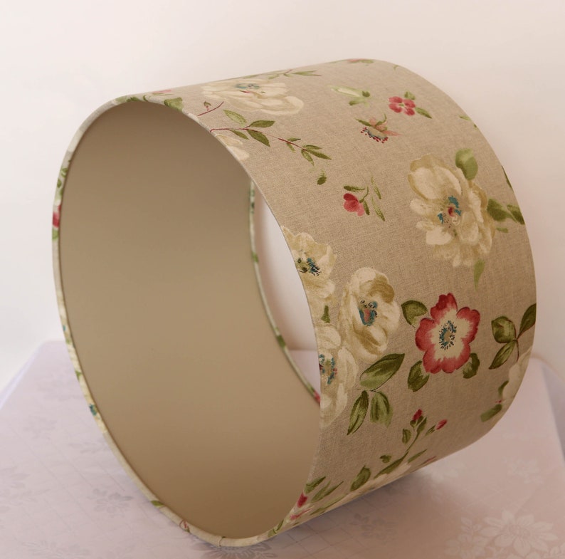 May include: A beige lampshade with a floral pattern featuring white, pink, and green flowers. The lampshade is made of fabric and has a white lining.