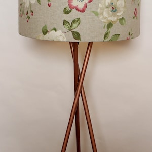 May include: A floor lamp with a tripod base made of copper colored metal. The lampshade is a light beige color with a floral pattern of white and pink flowers.