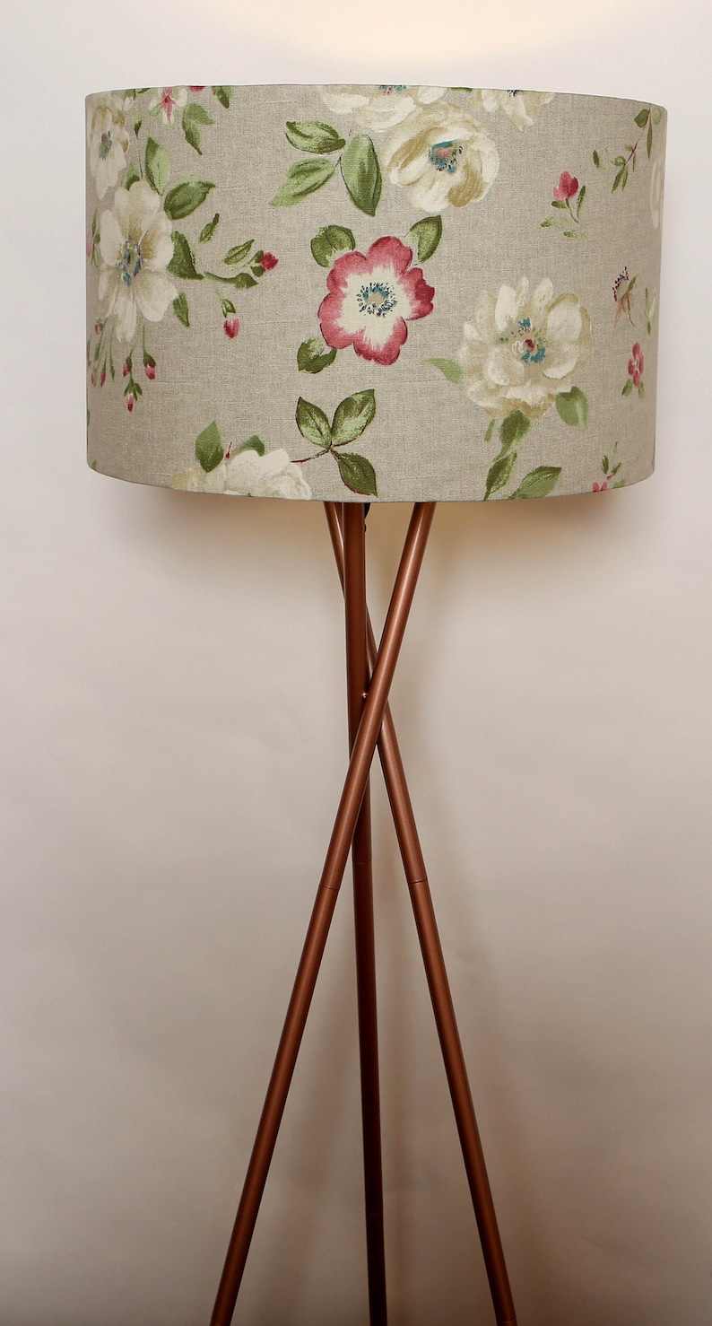 May include: A floor lamp with a tripod base made of copper colored metal. The lampshade is a light beige color with a floral pattern of white and pink flowers.