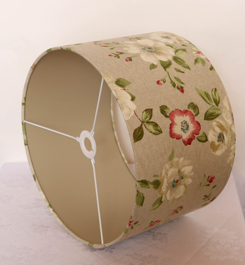 May include: A beige lampshade with a floral pattern featuring white, pink, and green flowers. The lampshade has a white fabric lining.