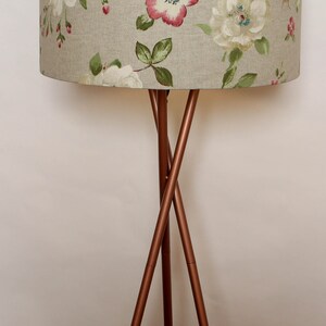 May include: A floor lamp with a tripod base made of copper colored metal. The lampshade is a light beige color with a floral pattern of white and pink flowers.