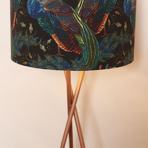 Może przedstawiać: Lampa stojąca z czarnym abażurem z motywem pawia. Abażur ma miedzianą podstawę z trzema nogami.