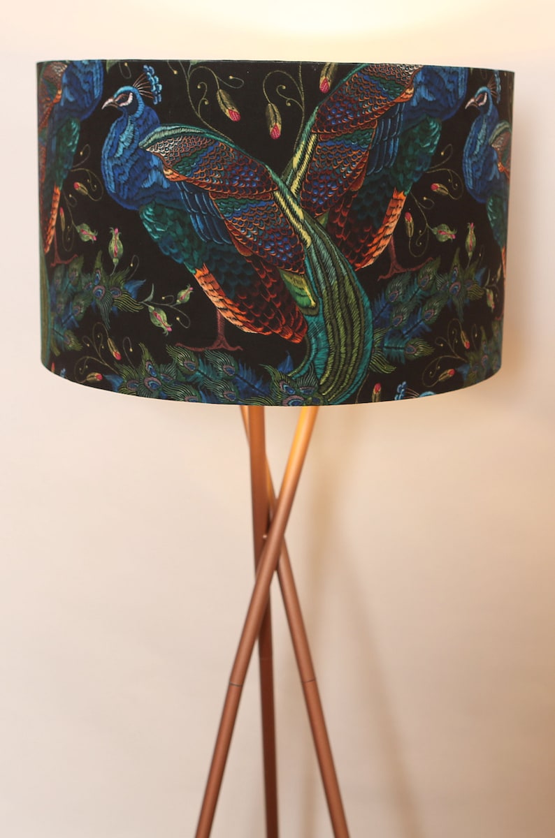 Może przedstawiać: Lampa stojąca z czarnym abażurem z motywem pawia. Abażur ma miedzianą podstawę z trzema nogami.