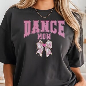 May include: A black t-shirt with the text "DANCE MOM" in pink letters and a pink bow with a heart in the center. The shirt is being worn by a person wearing gray shorts.