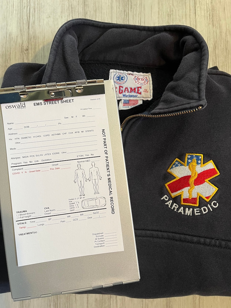 Puede incluir: Sudadera negra con cremallera con una estrella de la vida bordada en rojo, blanco y azul y la palabra "PARAMEDIC" en la parte delantera. La sudadera tiene una etiqueta en la parte delantera que dice "GAME Workwear" con una peque&ntilde;a bandera estadounidense.